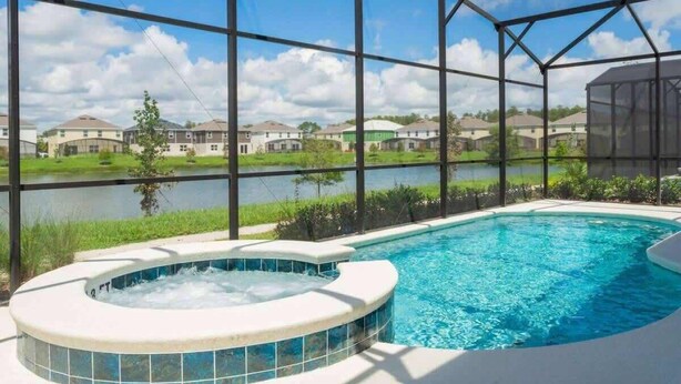 A swimming pool with a hot tub, surrounded by a covered area with a view of houses and a body of water.