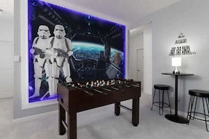 Una habitación con mesa de fútbol de mesa, decoración de pared con temática de Star Wars y una pequeña mesa con dos taburetes.