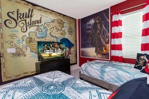 A room with a wall mural of Skull Island, a bed with a nautical-themed bedspread, a TV displaying a pirate-themed movie, and a window with red and white curtains.