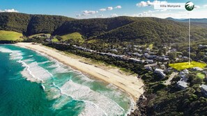 An aerial view of Villa Manyana, featuring a beachfront with clear turquoise waters, a sandy beach, and a residential area with houses.