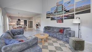 A modern living room with a grey sofa, a round coffee table, and a grey ottoman. There is a dining area with a table and chairs, and a kitchen in the background. - HUGE Home w/ Movie Room, Pool Table & Arcade Games (Indianapolis)