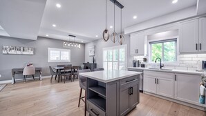 A modern kitchen with white cabinets, a central island, and a dining area with a gray table and chairs. - Cottage featuring Lake view balcony, private elevator and 3 en-suites! (Fort Erie)