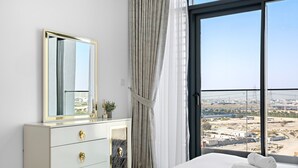 Habitación de hotel con un ventanal grande, una cama con ropa blanca, un aparador con tiradores dorados y un espejo.