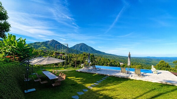 A pool area with lounge chairs and umbrellas, surrounded by lush greenery and mountains in the background. - Camaradvipa Retreat Munduk (Munduk)