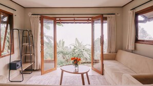 Une pièce comprenant un canapé, une table ornée d’un vase de fleurs, une échelle et une vue sur la verdure, visible à travers une porte vitrée.