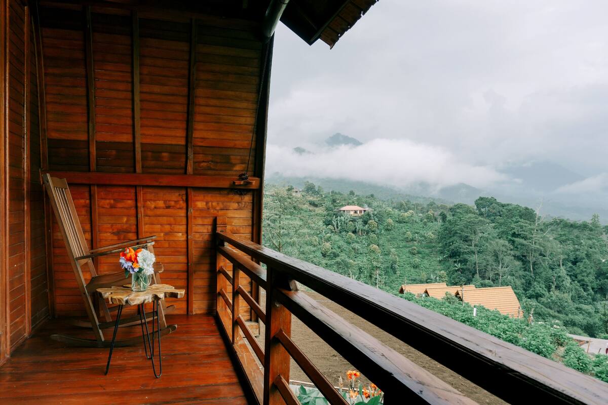 Ein Holzbalkon mit einem Stuhl und einem Blumenstrauß, mit Blick auf eine gebirgige Landschaft mit viel Grün und einigen Gebäuden.