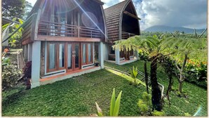 Une maison à deux étages dotée d’un balcon, entourée d’une végétation luxuriante et ayant une chaîne de montagnes en toile de fond.
