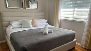 A neatly made bed with a grey bedspread and white pillows, a wooden floor, and a window with blinds.