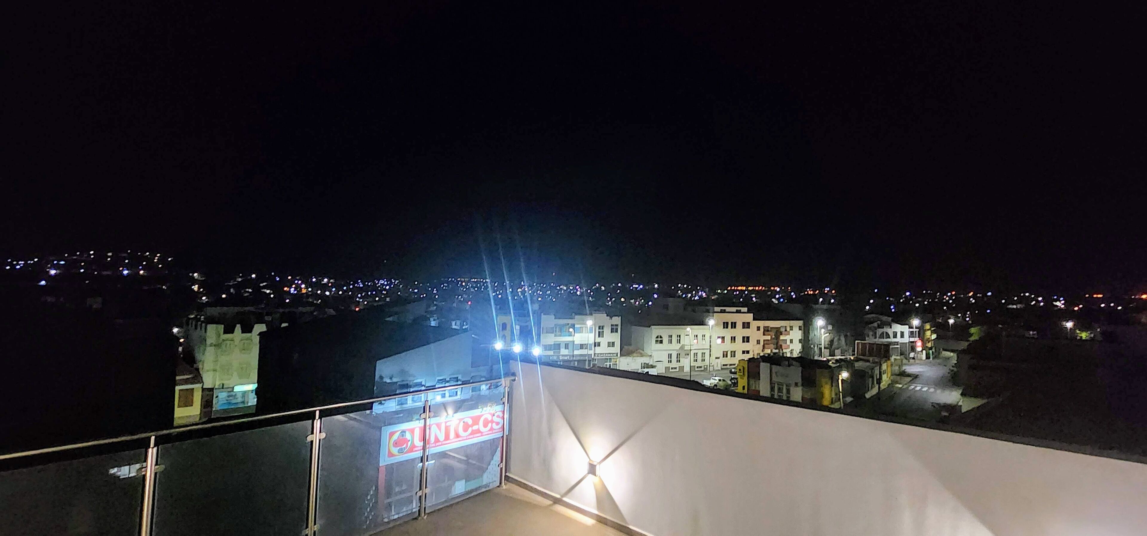A nighttime view from a balcony overlooking a cityscape with illuminated buildings and a visible sign for "UNTC-CS".