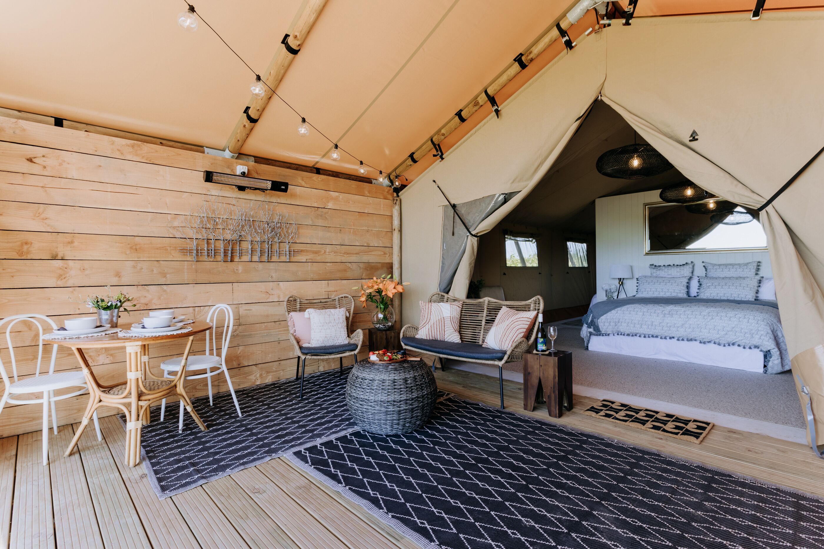 A tented accommodation with a wooden interior, a dining area with chairs and a table, and a bed with a view.