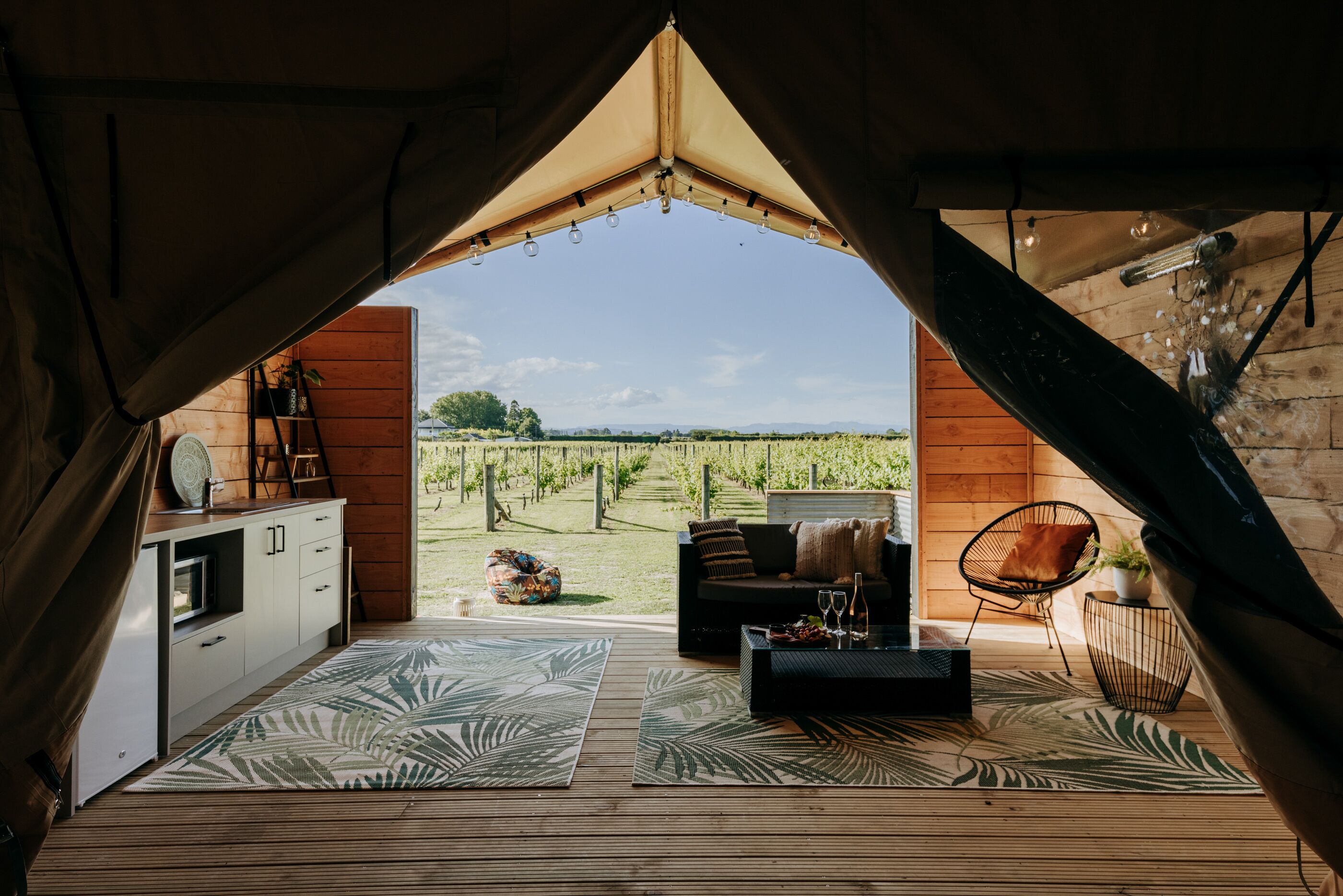 A tented accommodation with a kitchen area, seating, and a view of a vineyard.