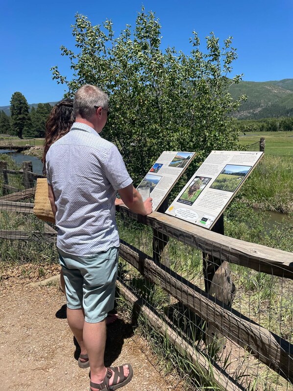 A man reading an informational sign at a park with a fenced area and a river in the background.