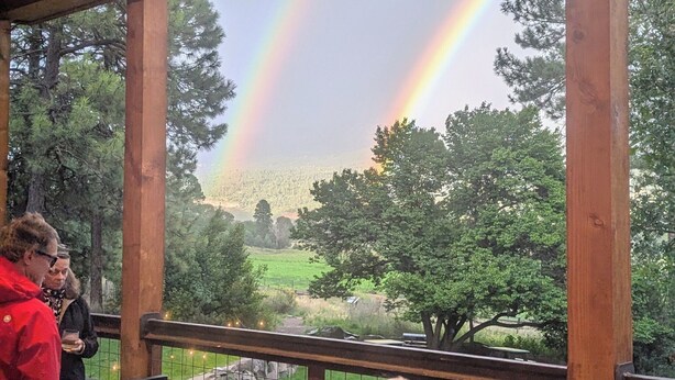 Ein überdachter Außenbereich mit Tischen und Stühlen, mit Blick auf eine Landschaft mit Bäumen und einem Regenbogen.