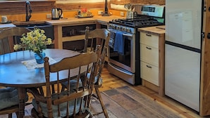 Ein rustikales Blockhaus-Interieur mit einem Holzespeisesaal, Stühlen, einer Küche mit Herd, Kühlschrank und einem Fenster mit Blick auf Grünflächen.