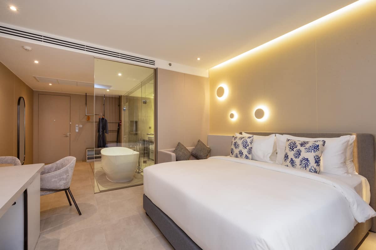 A modern hotel room with a large bed, a sitting area with a chair, a glass-enclosed shower, and a vanity with a mirror.