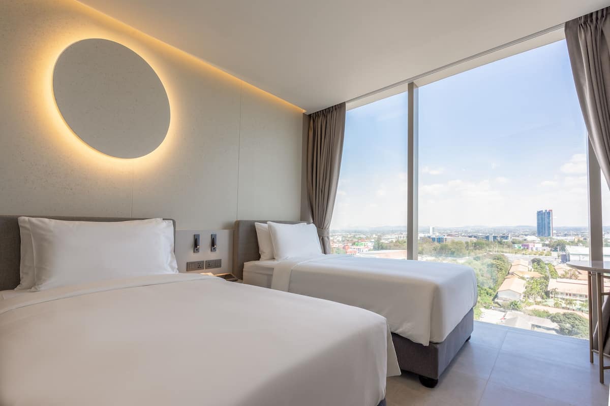 A hotel room with two beds, a circular light fixture on the wall, a large window with curtains, and a city view.