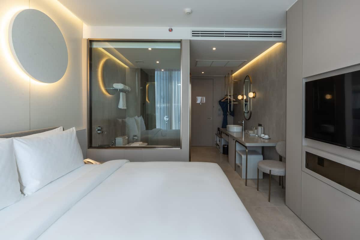 A modern hotel room with a large bed, a bathroom with a mirror, a TV, and a small seating area.