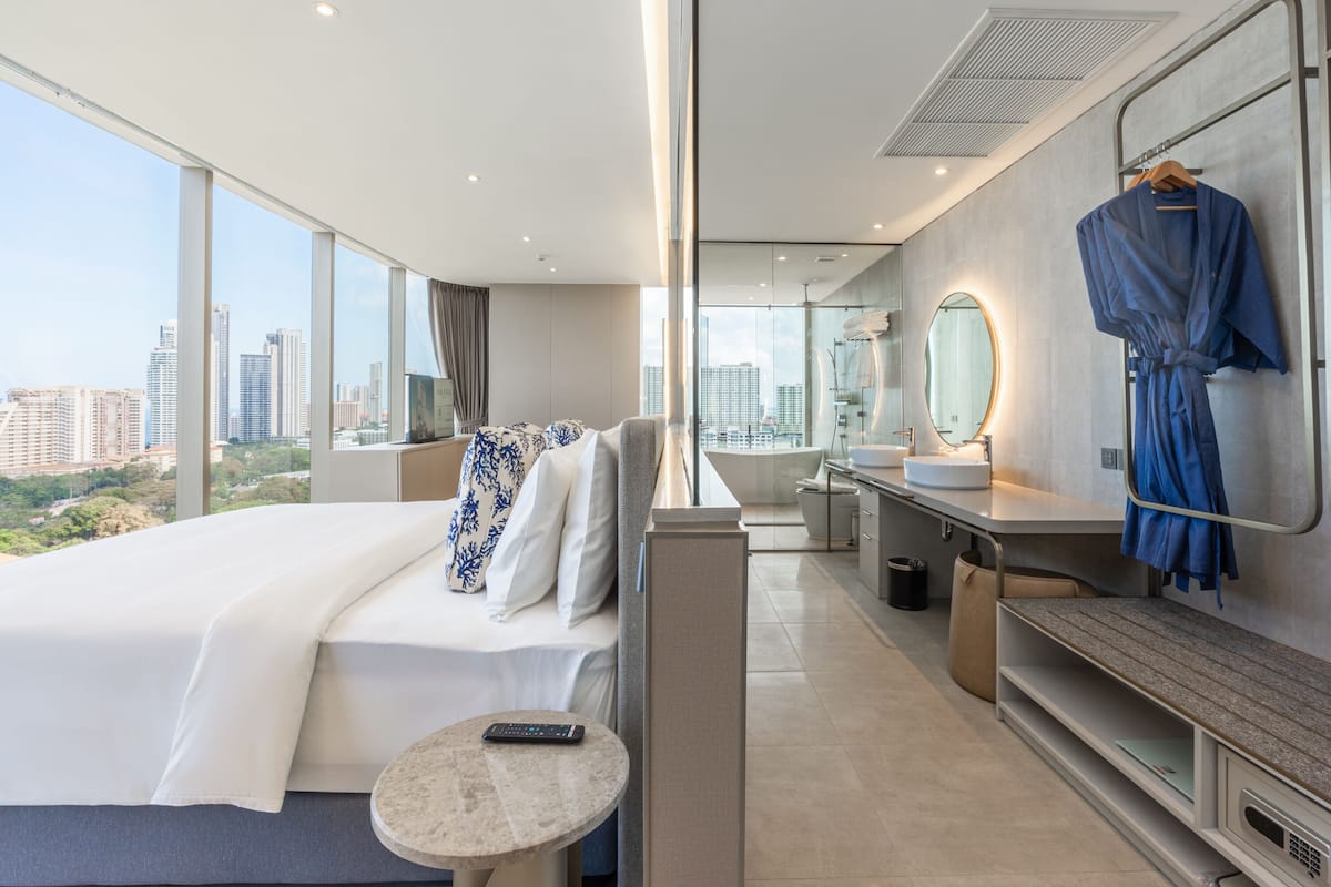 A modern hotel room with a large bed, a sitting area, a bathroom with a bathtub, and a city view.
