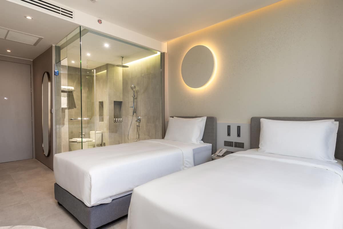 A modern hotel room with two beds, a glass-enclosed shower area, and a round mirror on the wall.