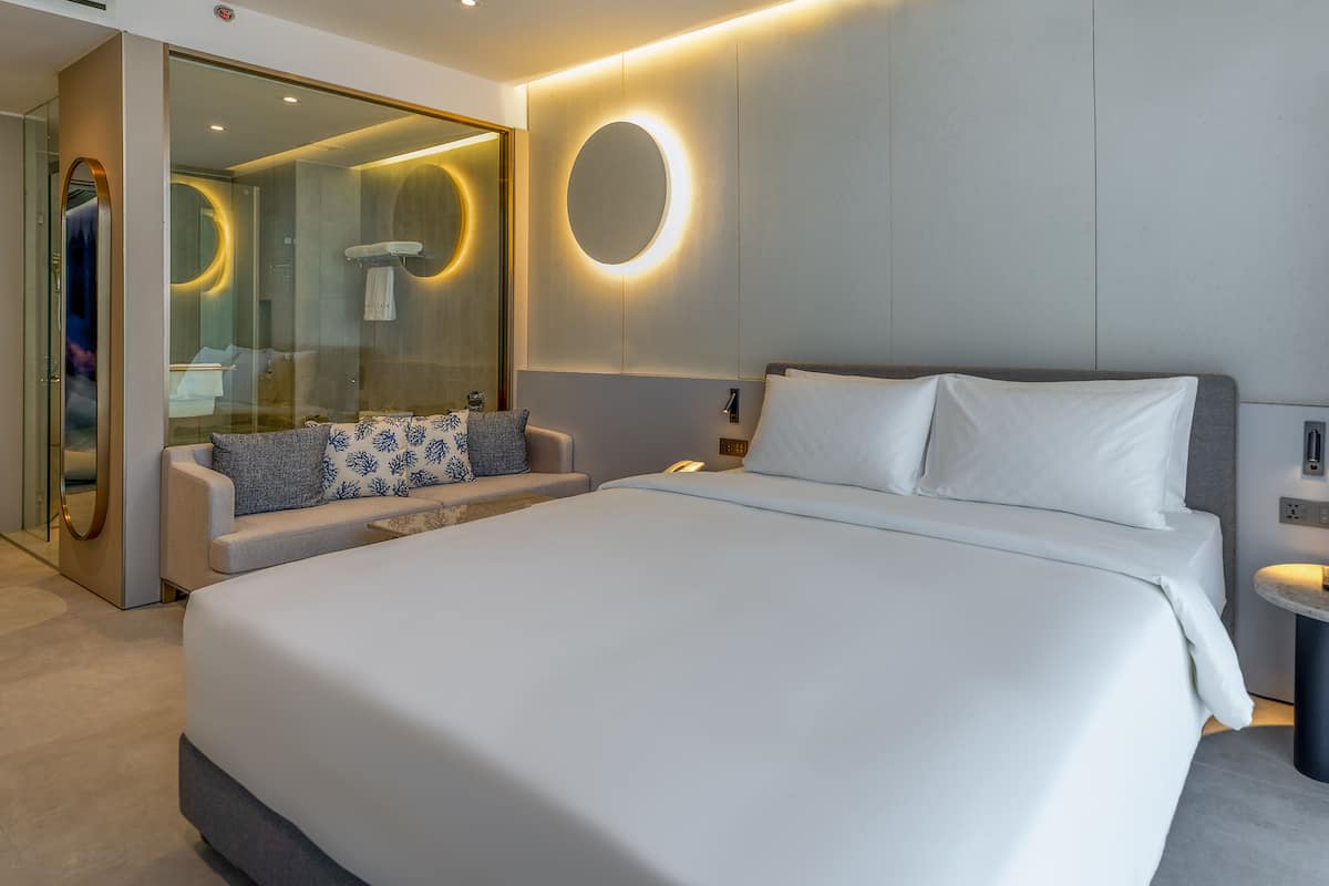 A modern hotel room with a large bed, a seating area with a sofa and pillows, a glass shower enclosure, and a round wall light.