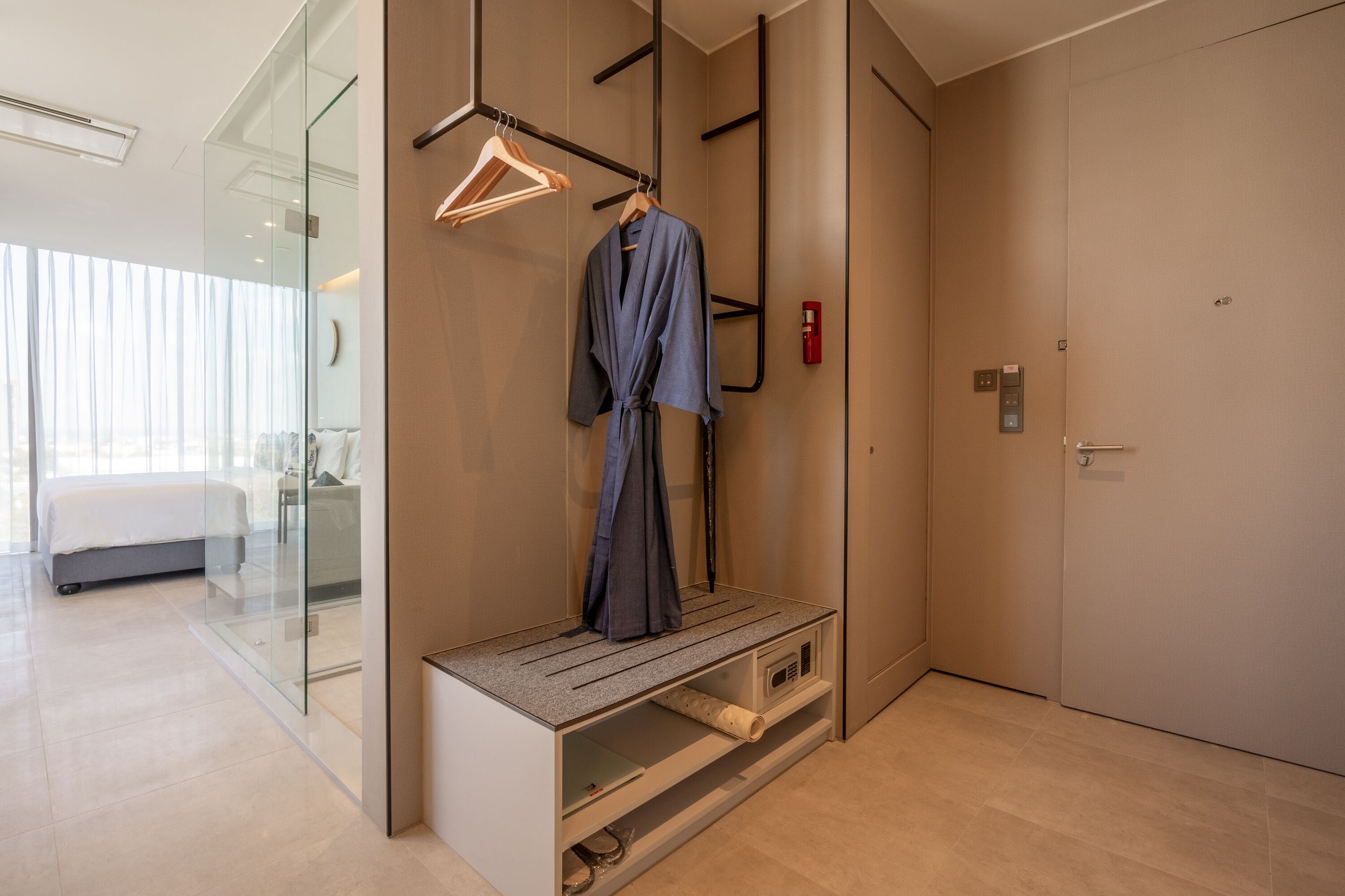 A modern hotel room with a bed, a bench with a robe, a coat rack, and a door.
