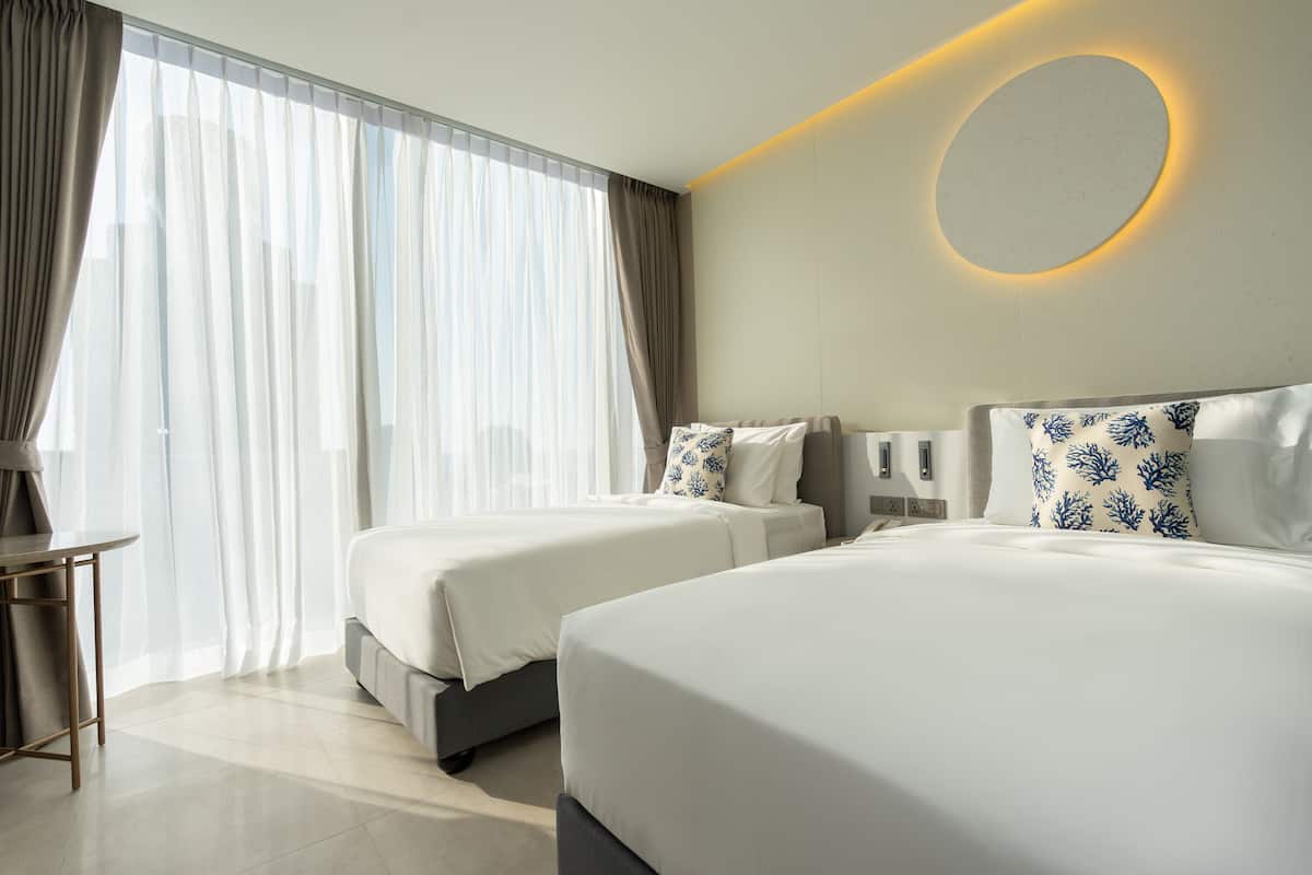 A hotel room with two beds, a desk, and a large window with sheer curtains.