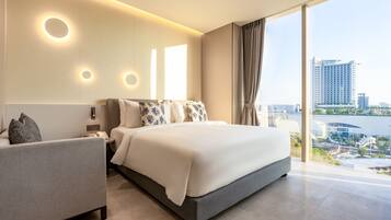 A modern hotel room with a large bed, a bedside table, and a view of a cityscape through large windows.