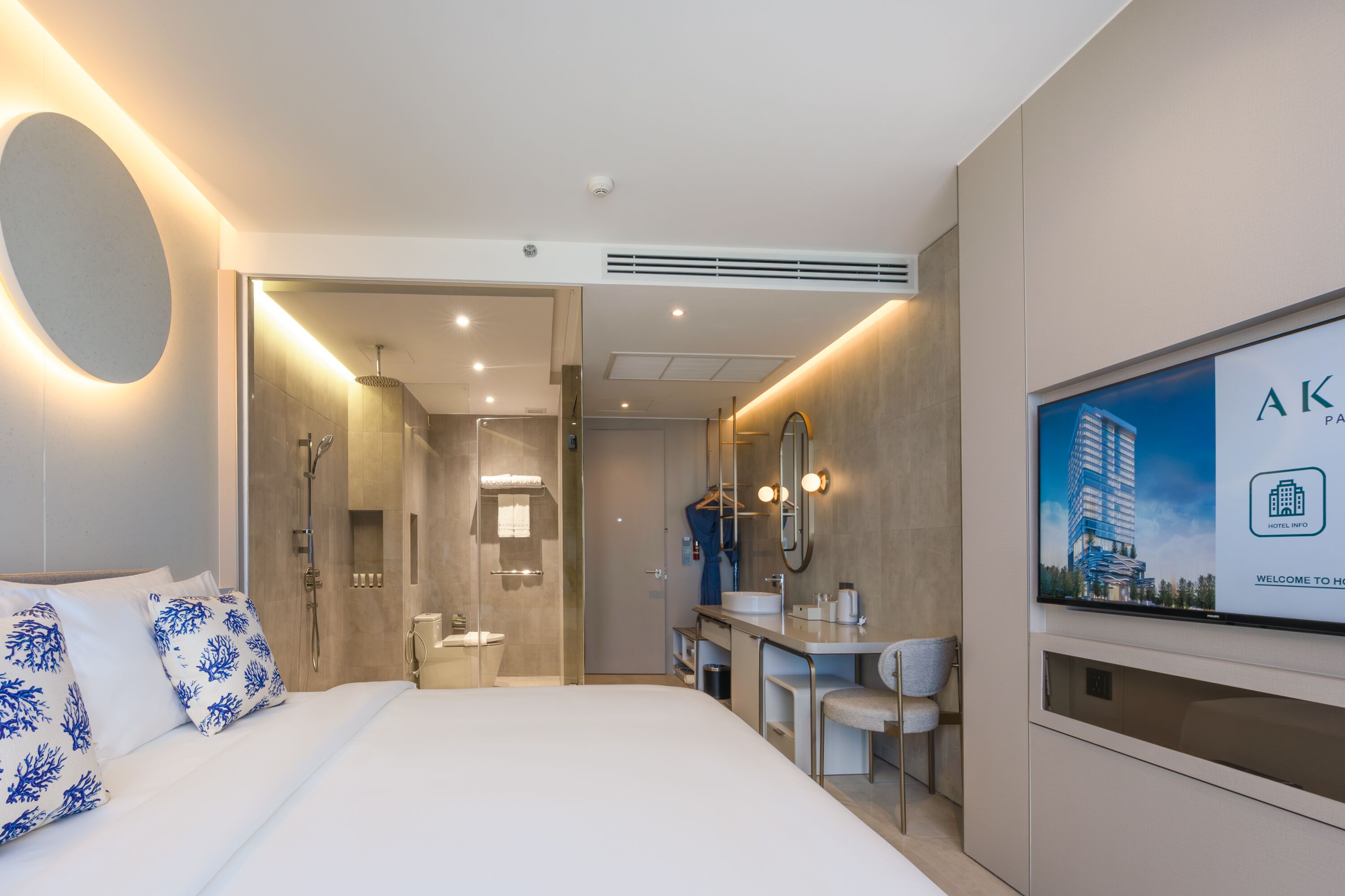 Una habitación de hotel moderna con una cama grande, un baño con ducha, un tocador con espejo y un televisor que muestra información del hotel.