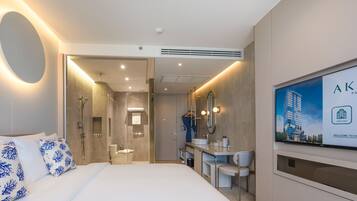 Una habitación de hotel moderna con una cama grande, un baño con ducha, un tocador con espejo y un televisor que muestra información del hotel.