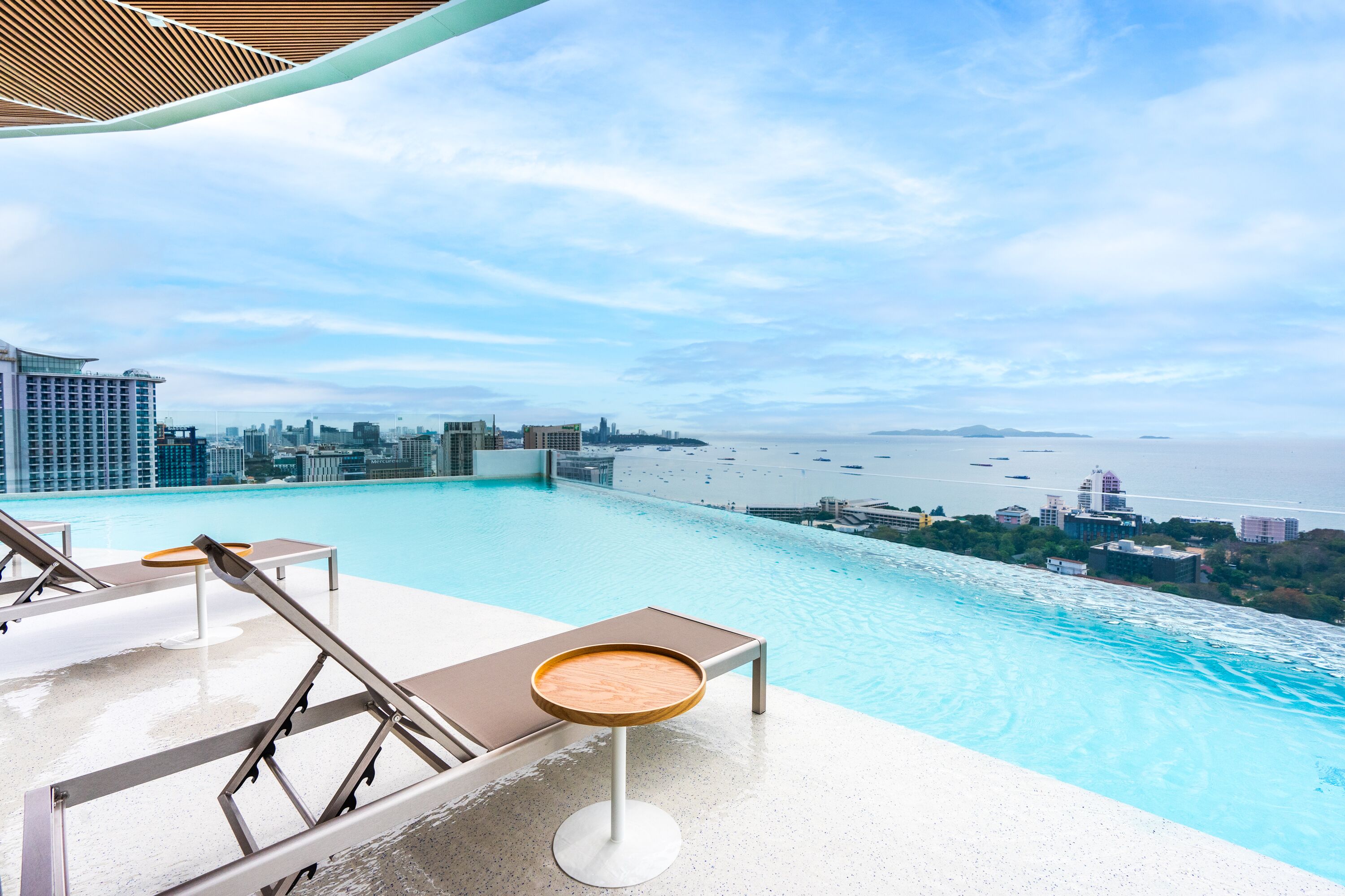 An infinity pool with lounge chairs and a table, overlooking a city skyline and the sea.