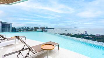 An infinity pool with lounge chairs and a table, overlooking a city skyline and the sea.