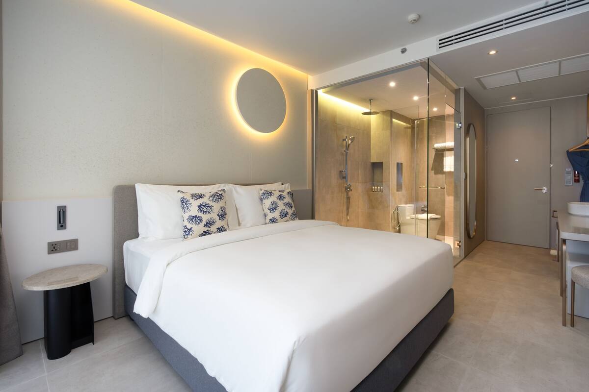 A modern hotel room with a large bed, a bedside table, a round mirror, a glass-enclosed shower, and a small bathroom with a sink and mirror.
