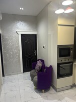 Una cocina moderna con horno empotrado y microondas, un sillón púrpura con un cojín decorativo y una puerta con marco negro.
