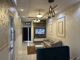 Una sala de estar moderna con un sofá color beige, un sillón azul, un televisor montado en la pared y un ventilador de techo.