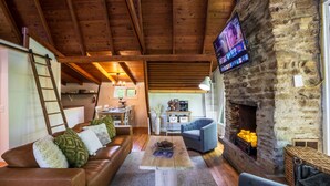 A cozy living room with a stone fireplace, wooden ceiling beams, a television mounted on the wall, and a comfortable seating area. - Lakefront with Lake Access•Hot Tub•Firepit•Pets (Rockbridge)