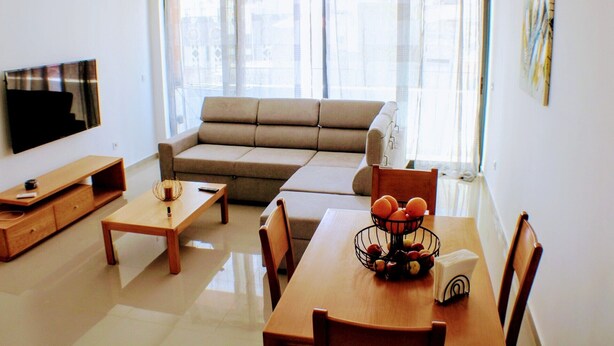 A living room with a sofa, a TV, a dining table with chairs, and a window with curtains. - Flamingo Plateau. 1A Apt on Rua Pedonal. Praia Central_ Near cafes & shops (Praia)