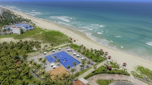 Vista aérea de un complejo turístico con piscina, sombrillas de playa y palmeras.