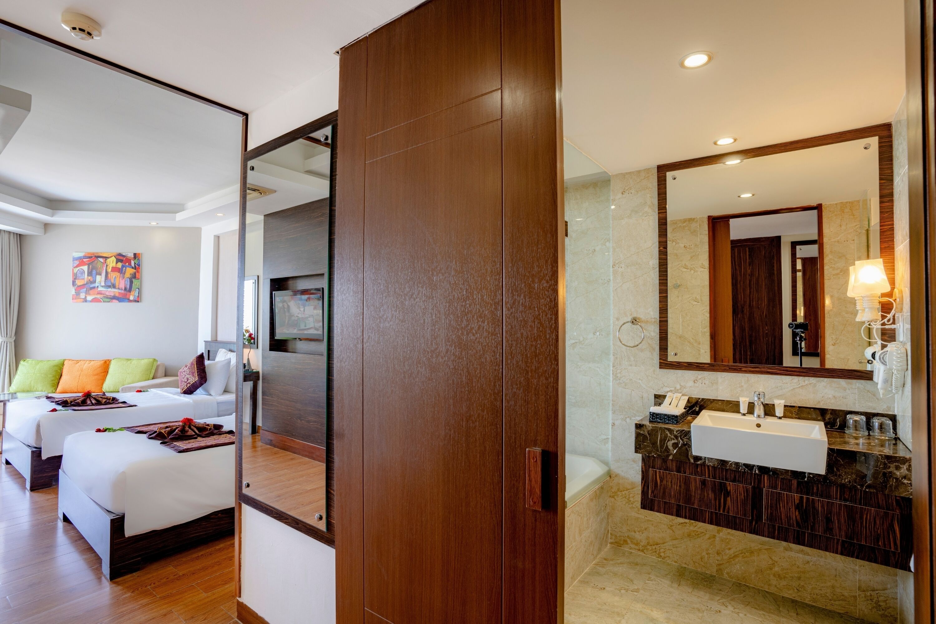 A hotel room with two beds, a large mirror, a bathroom with a sink and vanity, and a painting on the wall.