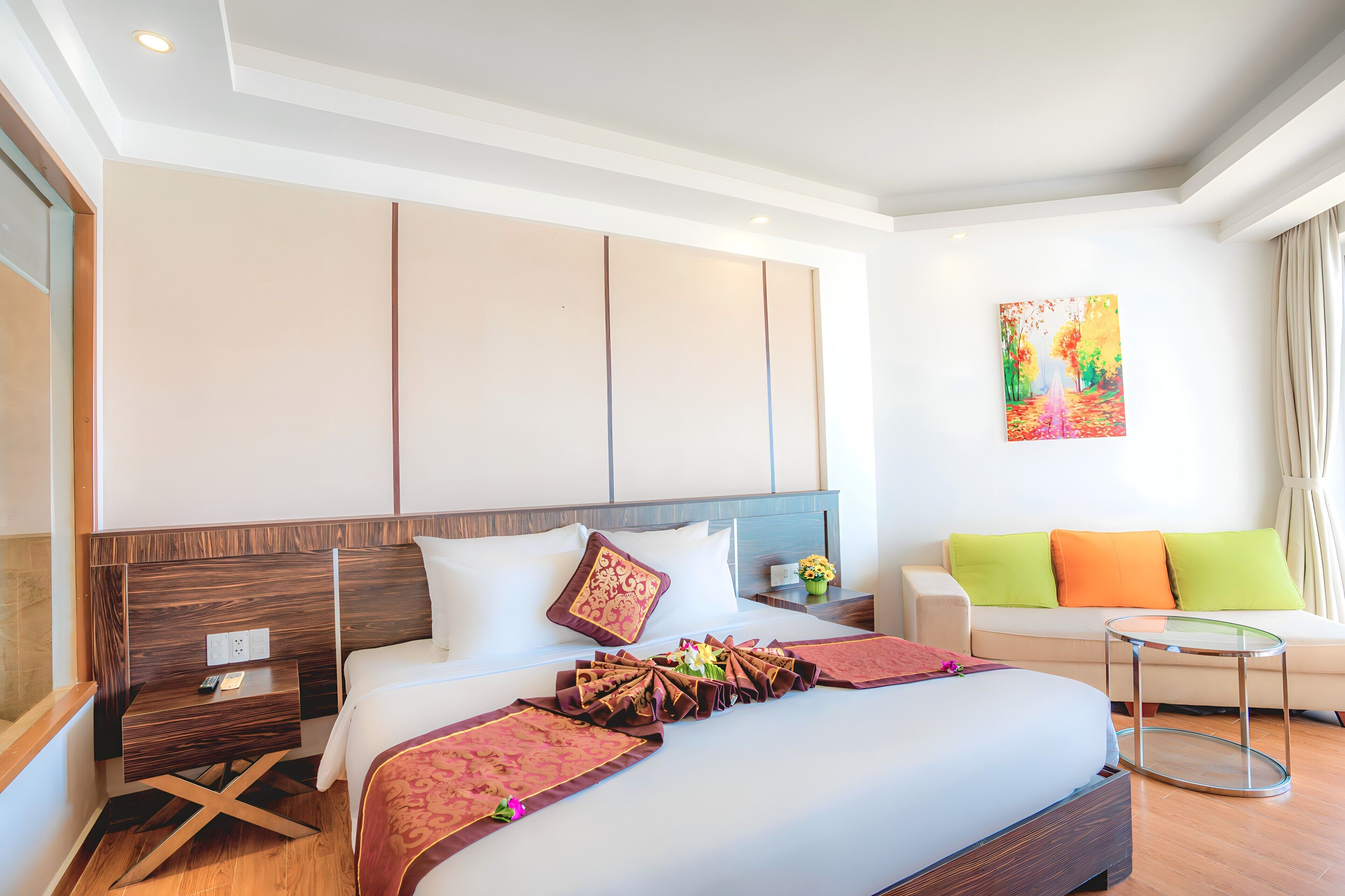 A hotel room with a large bed, a wooden nightstand, a glass side table, a sofa, and a colorful painting on the wall.