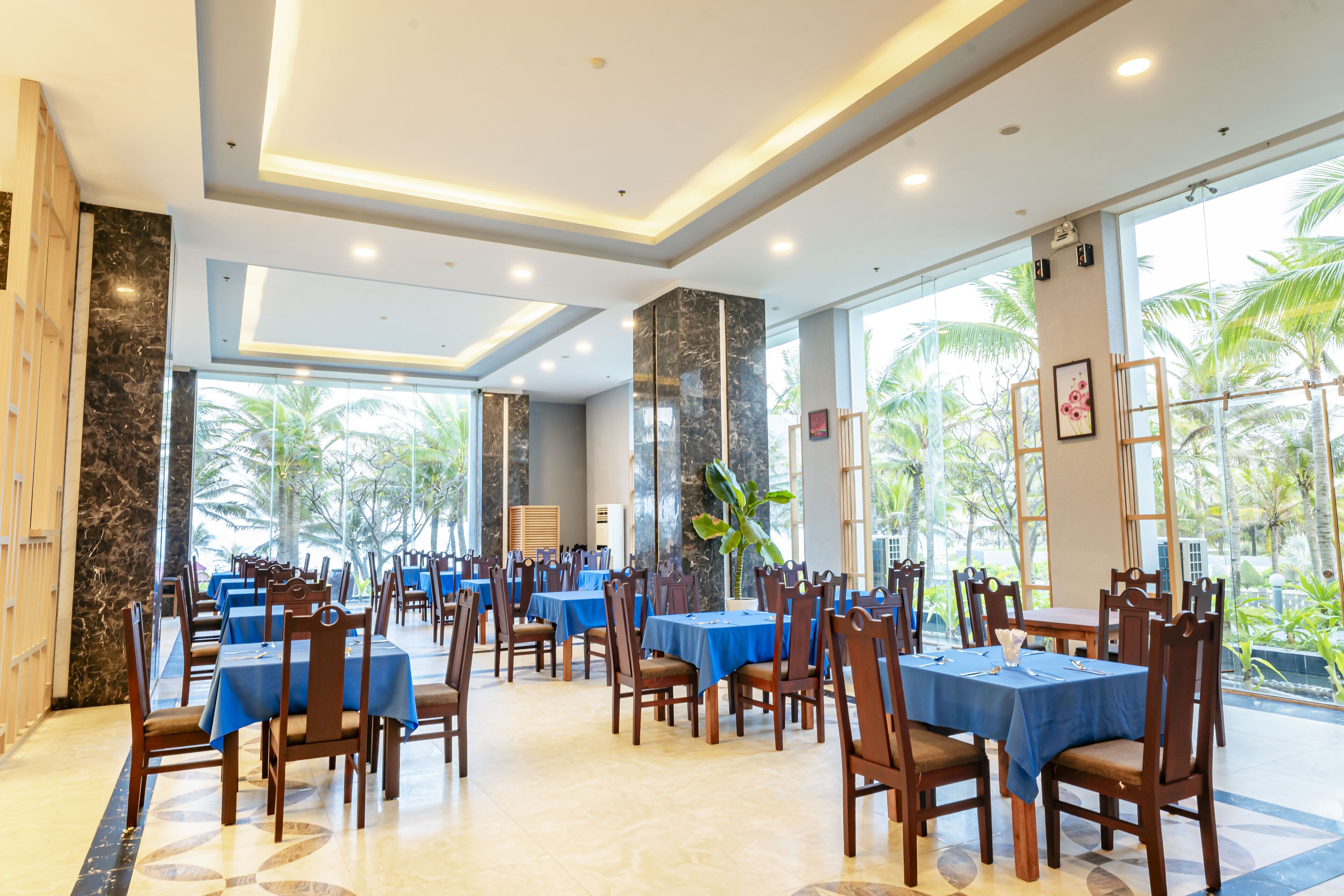 A spacious dining area with multiple tables and chairs, large windows with a view of palm trees, and a modern ceiling design.