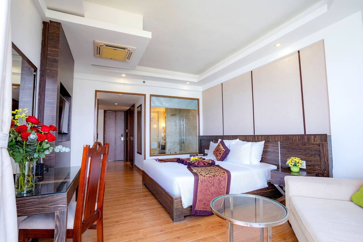 A modern hotel room with a large bed, a sitting area, a glass table, and a dining table with chairs.