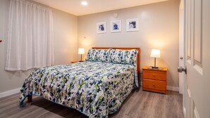 A bedroom with a floral bedspread, two bedside tables with lamps, a wooden nightstand, and a door. - Charming Apt_Open Green Space on Front to Relax and Sunbath (Hancock)
