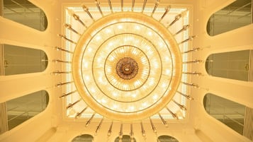 A grand, circular chandelier with multiple tiers and crystal accents, centered in a spacious atrium with high ceilings and arched windows.