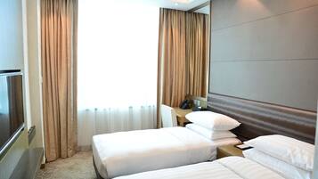 A hotel room with two beds, a desk, a chair, a TV, and a large window with curtains.