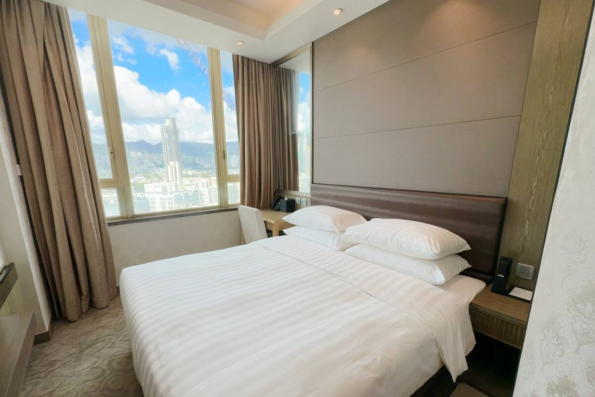 Ein Hotelzimmer mit einem großen Bett, einem Nachttisch, einem Schreibtisch und Blick auf eine Stadtlandschaft durch das Fenster.