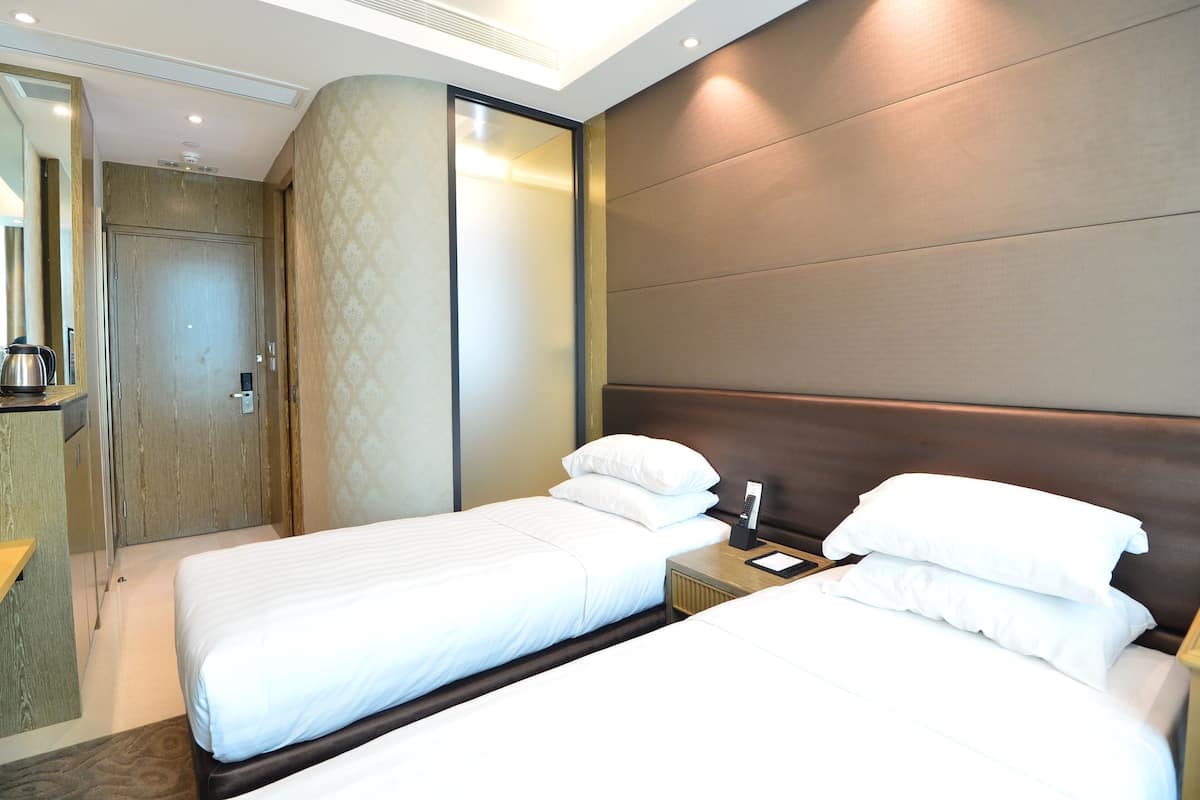 Ein Hotelzimmer mit zwei Betten, einem hölzernen Kopfteil, einem Nachttisch mit Lampe sowie einem Badezimmer mit Dusche und Toilette.