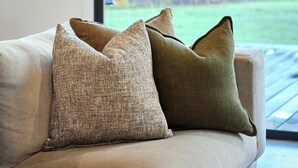 A beige sofa with two pillows, a framed picture on the wall, and a view of trees and grass through a window. - Escape to a unique house located in the world-renowned surf spot of Bells Beach! (Bells Beach)