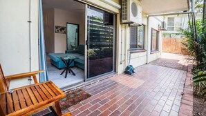 Une terrasse avec une table et des chaises en bois, un canapé bleu et une porte-fenêtre coulissante donnant sur un espace intérieur agrémenté d’un mur végétal et d’un tableau encadré.