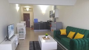 Een woonkamer met een groene bank, een witte salontafel, een televisie en een keuken op de achtergrond.