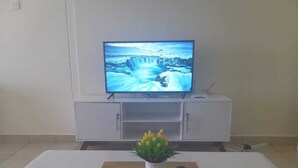Een woonkamer met een flatscreen televisie op een wit kastje, een witte salontafel met een plant en een afstandsbediening, en een beige bank.